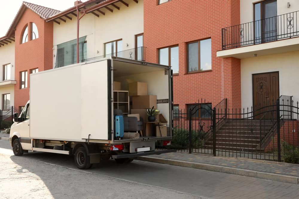 A moving truck is parked on the street in front of a modern, multi-story brick townhouse. The truck's back door is open, revealing various packed boxes, furniture, and household items inside. With the help of professional residential moving services, a flight of stairs leads up to the main entrance of the house.