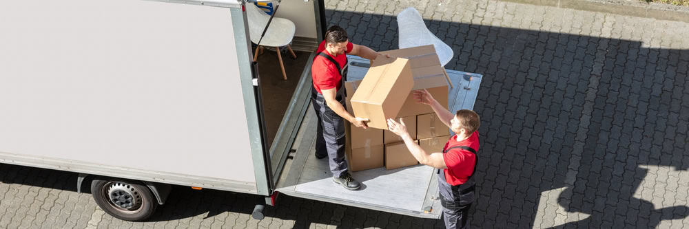 Two movers in red shirts and black overalls are unloading large cardboard boxes from the back of a moving truck. Specializing in commercial moving, one man stands on the truck’s lift gate, handing a stack of boxes to the other man, who is stationed on the ground.