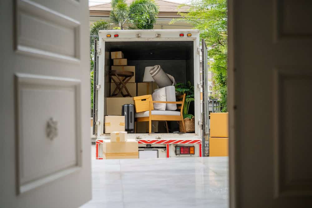 A moving truck filled with various household items, including boxes, chairs, plants, and a rolled-up rug, is parked outside an open door. The view is from inside a house, looking out toward the truck—an evident sign of efficient furniture moving in action.