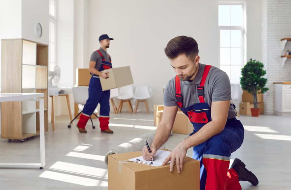 Two movers in uniforms are working in a bright room. One is kneeling and writing on a clipboard placed on a cardboard box, while the other is carrying another box. The room, set for Residential Moving, has large windows, white walls, and minimal furniture.