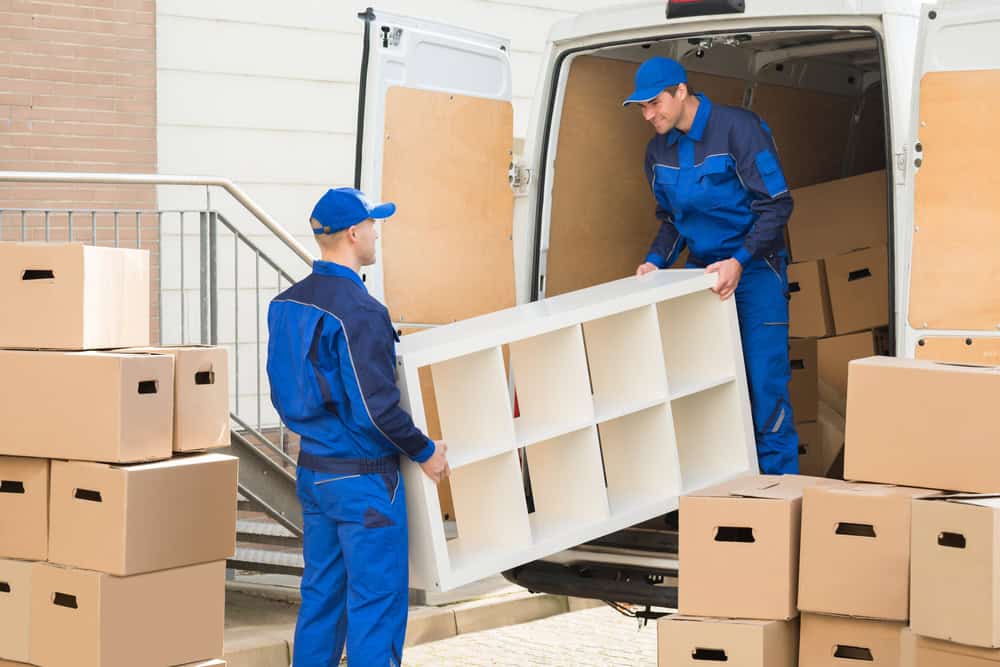 Two movers in blue uniforms, offering professional moving services, are unloading a white shelving unit from a van filled with packed brown boxes. One mover is inside the van while the other is outside, both holding the shelving unit. Several boxes are stacked on the ground next to the van.