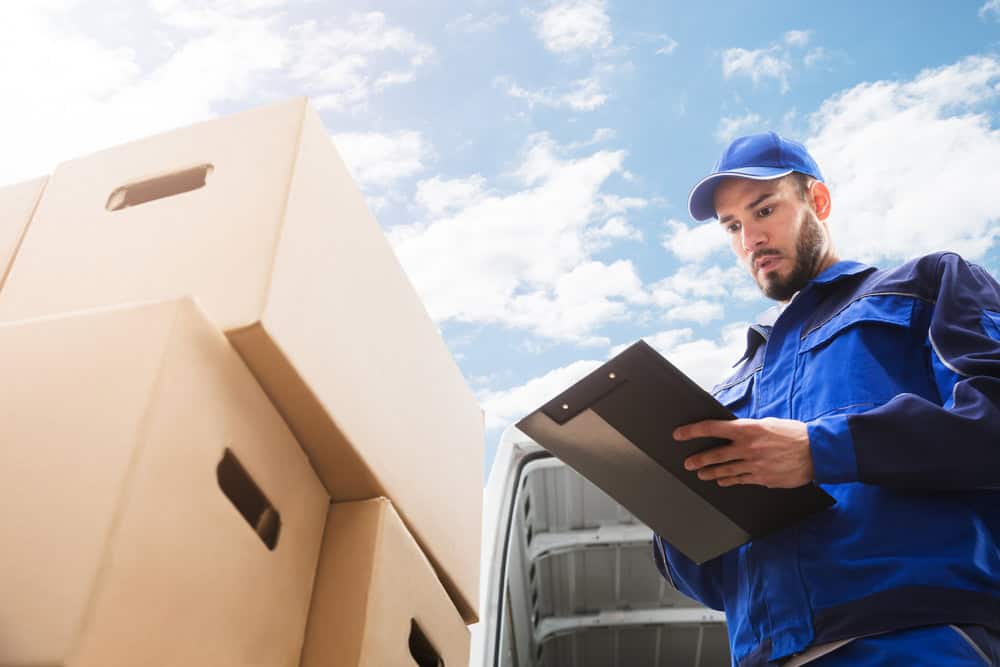 A delivery worker in a blue uniform and cap stands next to stacks of cardboard boxes, holding a clipboard. The sky is partly cloudy in the background, and the back of a delivery van is open, indicating he is likely preparing for or checking a commercial moving service.