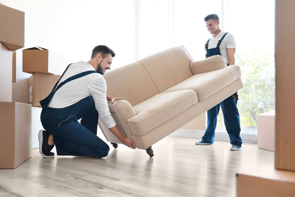 Two men in blue overalls and white shirts are lifting a beige sofa inside a bright room with light wooden flooring. Several cardboard boxes around them signal a residential moving process.