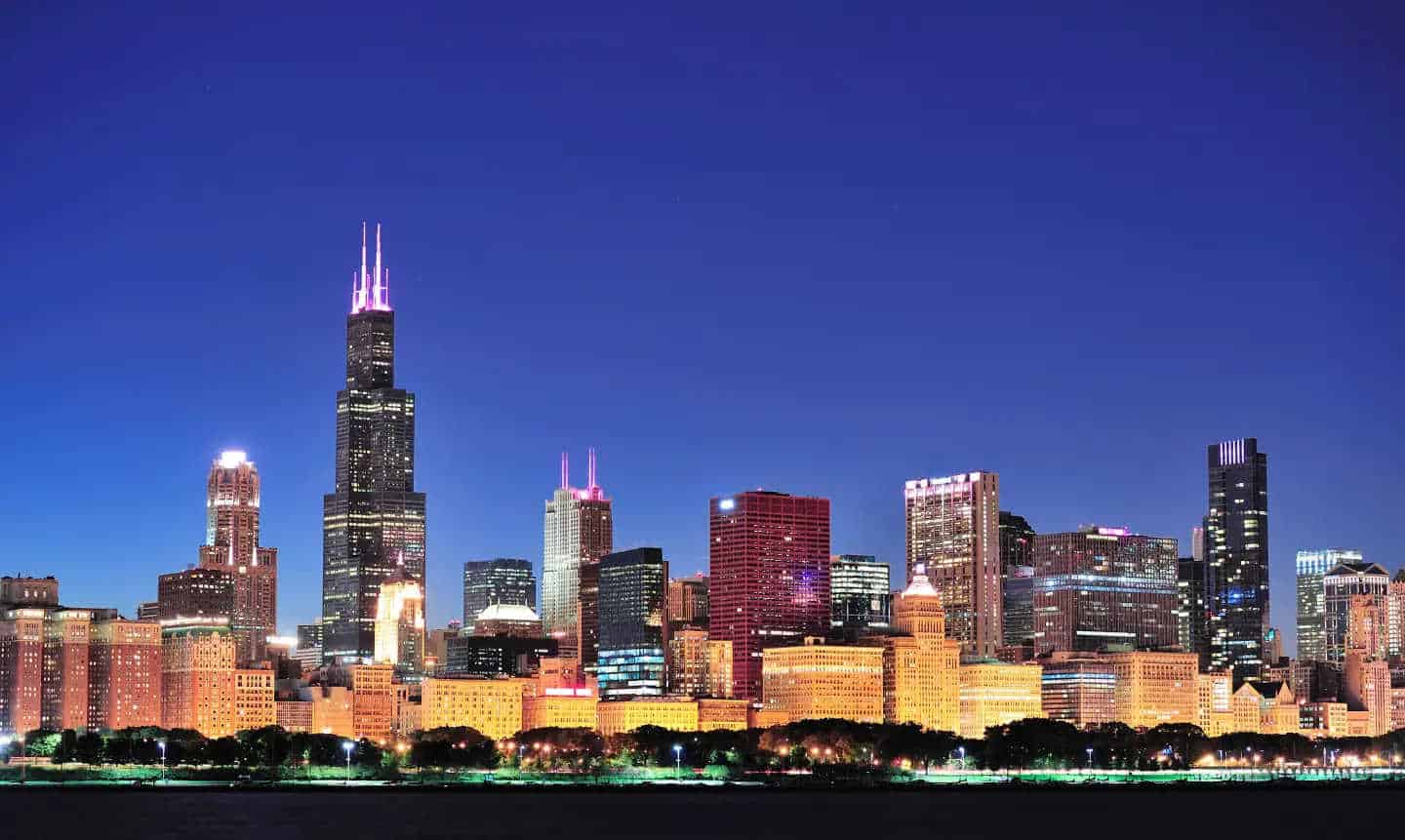 A nighttime view of a city skyline featuring various lit-up skyscrapers against a clear, dark blue sky. The tallest building has prominent twin antennas at the top, illuminated in pink. The foreground features silhouettes of trees and buildings softly aglow with lights, highlighting bustling moving services below.