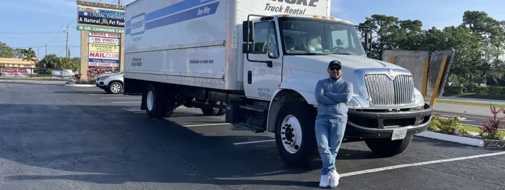 A person leans against a large white moving truck with their arms crossed, wearing a gray hoodie, blue jeans, and a baseball cap. The truck, advertising professional Moving Services, is parked in a lot with a commercial sign featuring multiple store names visible in the background on a sunny day.