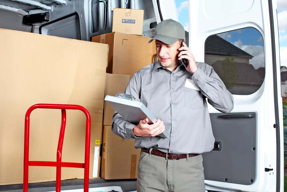 A delivery worker in a gray uniform and cap speaks on a phone while holding a clipboard. He stands inside a van filled with cardboard boxes, preparing for the next Commercial Moving job. A red hand truck is visible in the foreground.