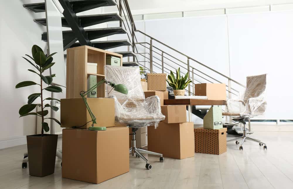 An office space with various pieces of furniture and equipment packed in cardboard boxes, indicating a recent commercial moving operation. Two office chairs wrapped in plastic, a desk with a green lamp, and plants are also in view. A staircase with black railings is in the background.