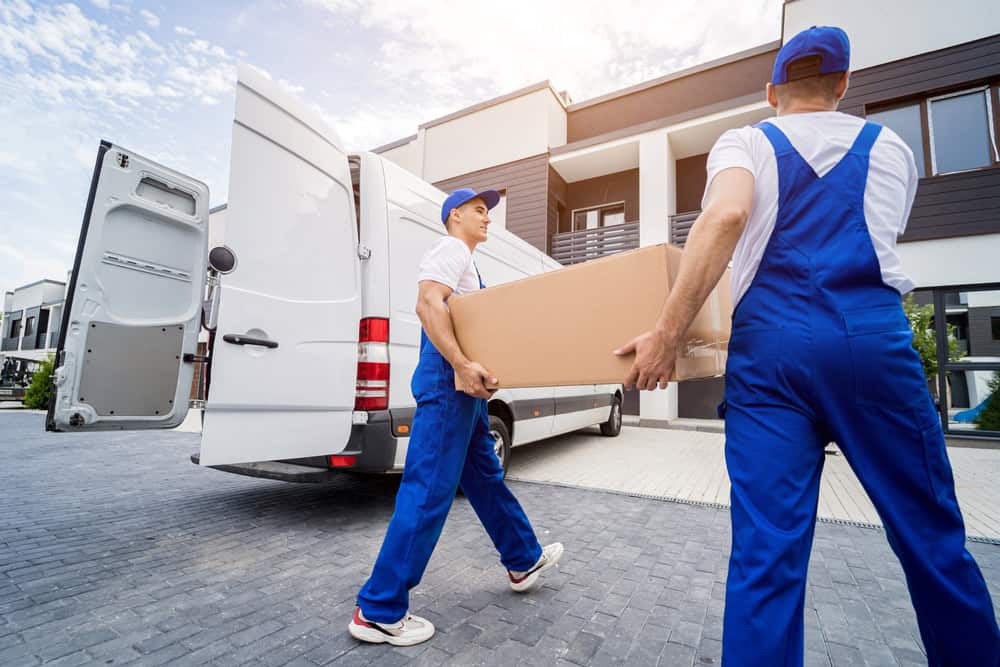 Two movers in blue overalls and white t-shirts are carrying a large cardboard box from a white van towards a modern townhouse. The van's rear doors are open, signifying ongoing moving services. The day appears to be bright and sunny, perfect for furniture moving or even commercial moving tasks.