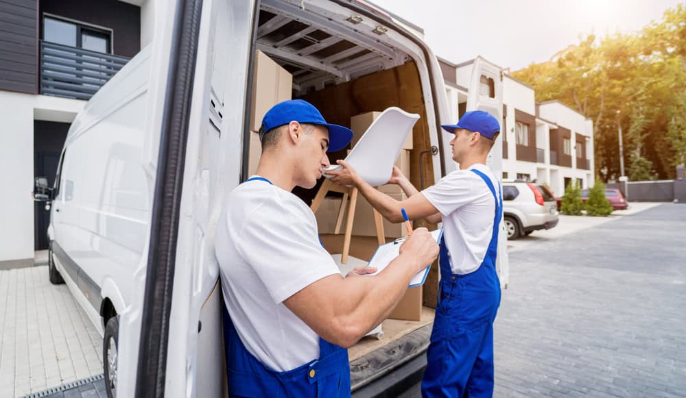 Two movers in blue uniforms are working near an open moving van in a residential area. One is checking a clipboard, while the other is lifting a white chair into the van. Various packed boxes are visible inside the van, highlighting their expertise in furniture moving, with modern buildings in the background.
