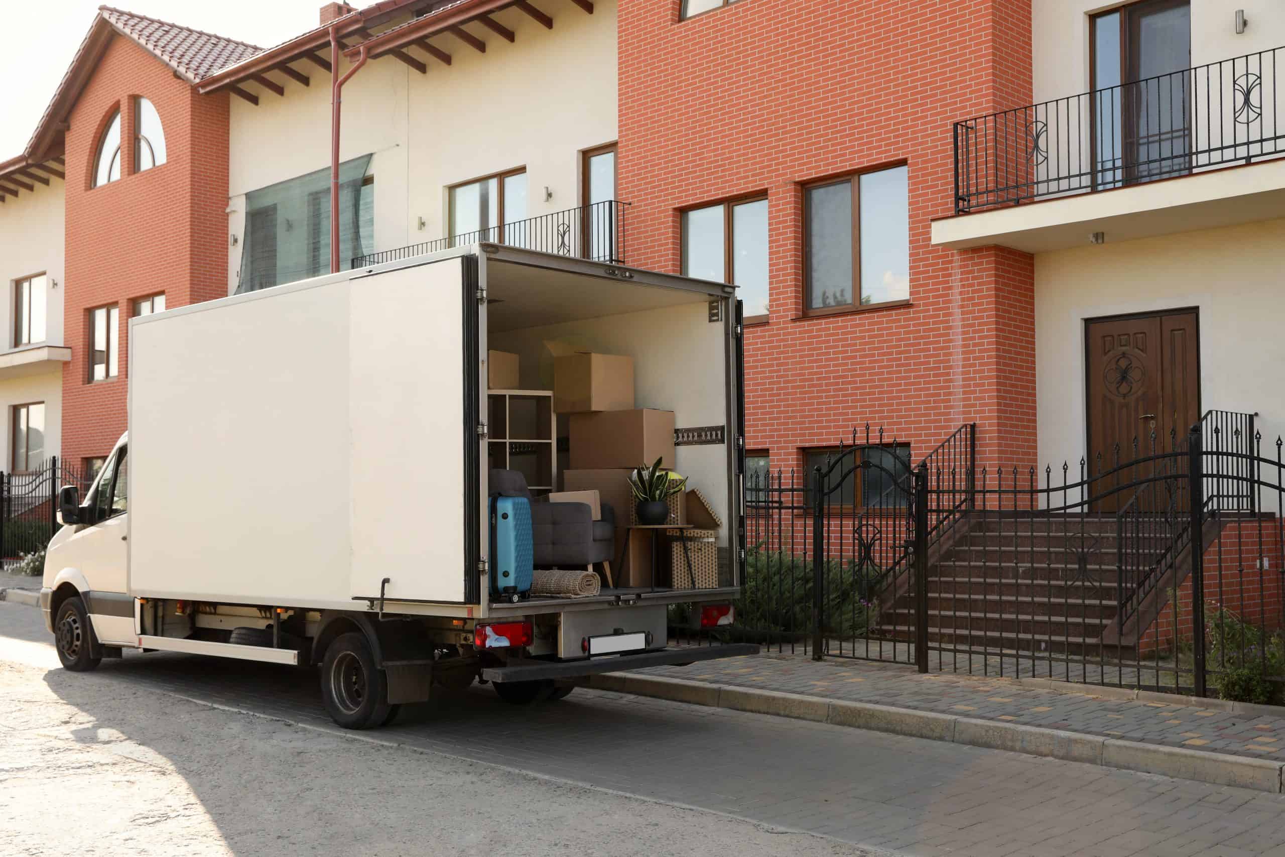 A moving truck is parked outside a modern brick apartment building. The truck's back door is open, revealing packed boxes, a rolled-up rug, a suitcase, and a potted plant inside. This scene suggests that residential moving services are in progress and someone is in the process of moving in or out.