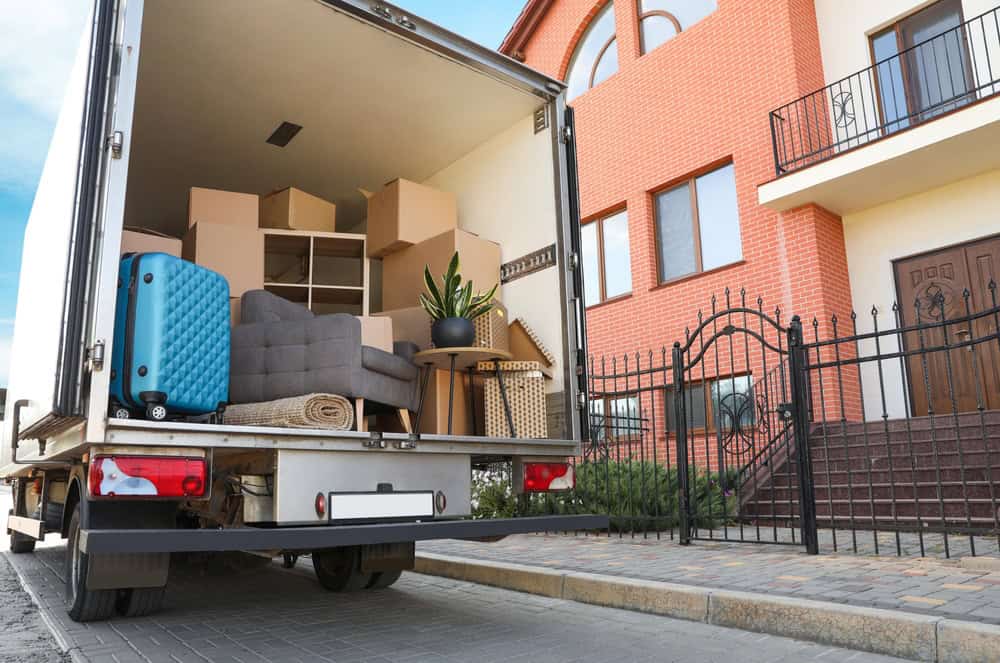 A moving truck is open, revealing various items packed inside for a residential moving job. Items include a turquoise suitcase, a grey sofa, a rolled-up rug, and a potted plant. The truck is parked in front of a red brick house with a black metal fence and double door entrance.