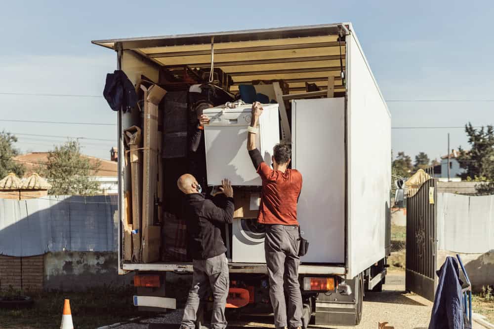 Two men loading household items into a large white moving truck, clearly part of a professional moving service. One man is holding a white appliance while the other assists, both standing at the open back of the truck. The background displays a typical residential area with fences and houses.