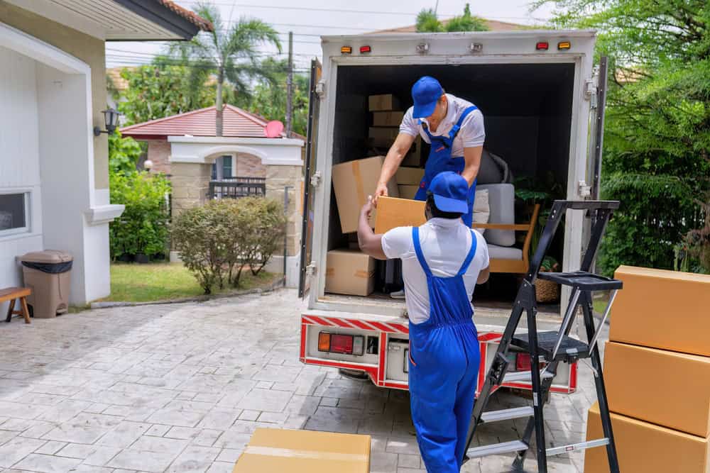 Two movers in blue uniforms are unloading boxes as part of their residential moving services in front of a house. One mover hands a box down to the other while additional boxes and a ladder are nearby. The surrounding area has greenery, with other houses visible in the background.