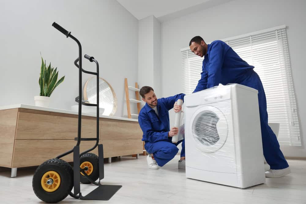 Two workers in blue uniforms are unpacking a wrapped washing machine in a bright, modern room. A dolly with yellow wheels is standing nearby. The room, serviced by residential moving experts, features light wood furniture, a large mirror, and a plant. White blinds cover the windows.