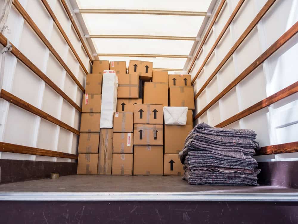 A loaded moving truck with stacked cardboard boxes of various sizes and a few white cushioned items, ideal for residential moving. Several folded moving blankets are neatly placed on the floor of the truck. The boxes have arrows indicating the upright position.