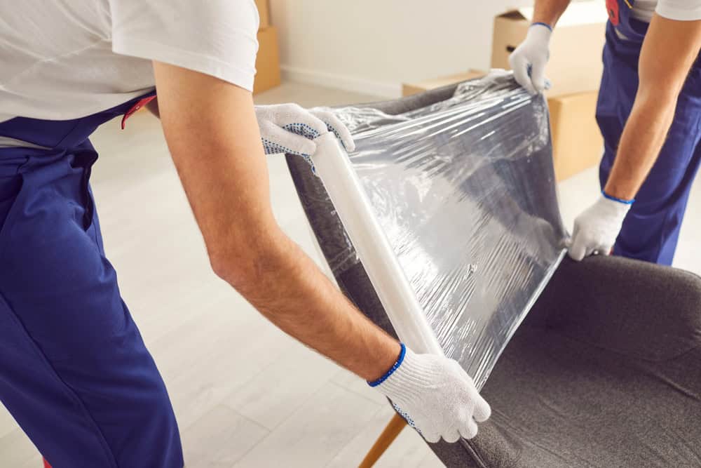 Two individuals wearing gloves and blue pants are wrapping a gray chair with plastic film, presumably preparing it for moving or storage. Brown cardboard boxes are visible in the background, highlighting their expertise in commercial moving services.