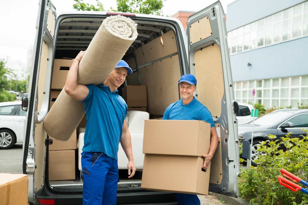 Two movers in blue uniforms are unloading a van. One man, smiling at the camera, carries a rolled-up carpet on his shoulder while the other holds a cardboard box. The van's back doors are open, revealing more boxes and items inside—a snapshot of efficient residential moving services in action.