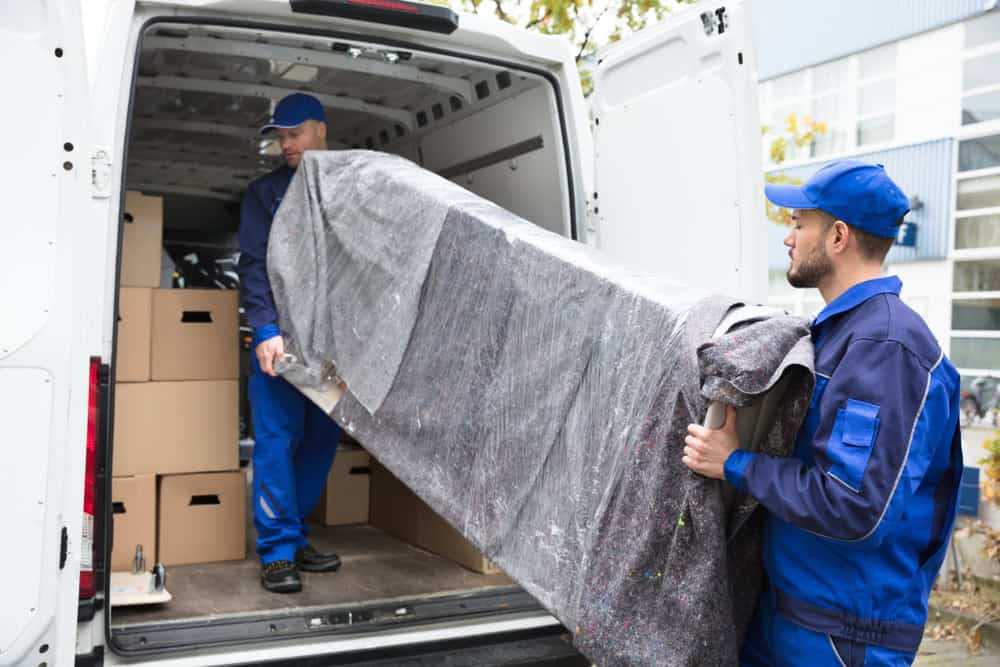 Two movers in blue uniforms are loading a wrapped piece of furniture into a white moving van marked with "Commercial Moving Services." The interior of the van is visible, showing several cardboard boxes stacked inside. One mover is inside the van, while the other stands outside holding the furniture.
