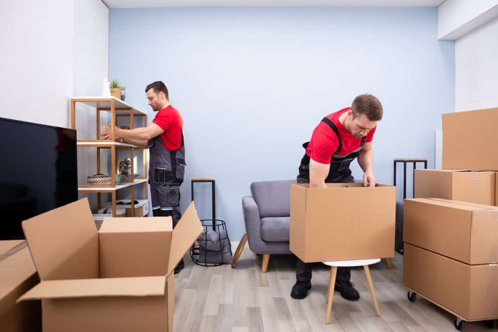 Two movers in red shirts and overalls are unpacking boxes in a living room. One is arranging items on a shelf, while the other is opening a box on a small table. The room has a sofa and several cardboard boxes.